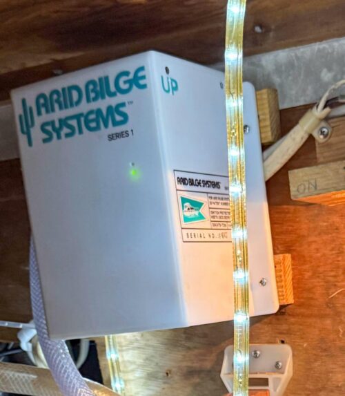 Arid Bilge Systems Series 1 white enclosure with turquoise cactus logo mounted inside a boat, with connected hoses and an illuminated indicator.