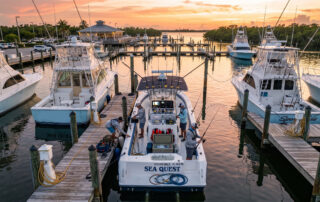 Sunset over a marina with several white boats docked at piers and calm water reflecting the orange sky, center boat named Sea Quest in view.