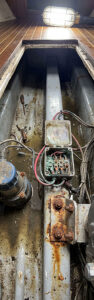 Maintenance access pit with a rusty electrical junction box, exposed wiring, and a dirty water-filled trench, illuminated by a ceiling work light above.