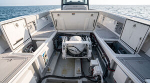 Open boat engine bay with a central white circular component, surrounded by black and colored hoses and storage lockers, ocean in the background.