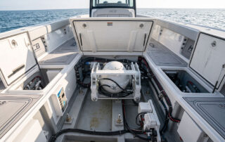 Open boat engine bay with a central white circular component, surrounded by black and colored hoses and storage lockers, ocean in the background.