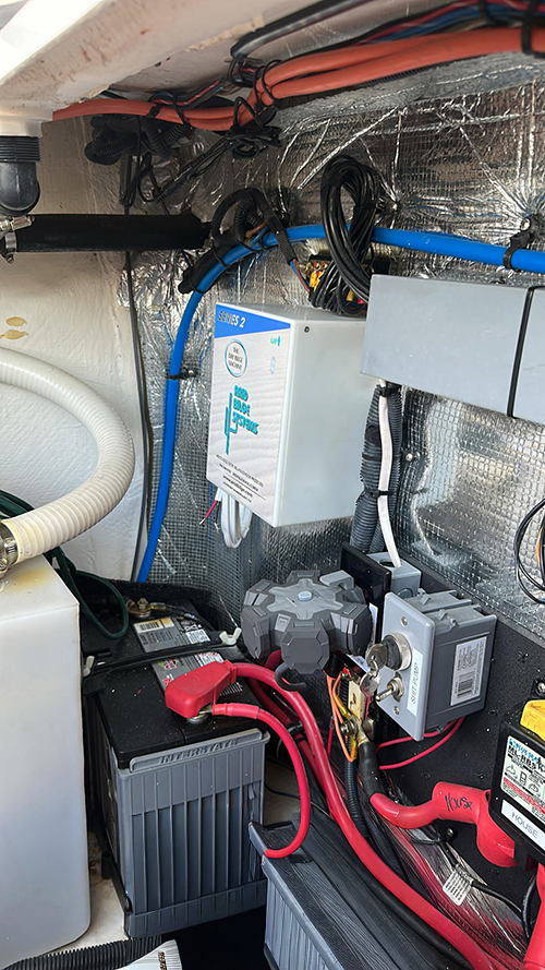 Utility space with a car battery and red/black cables, a white control box, blue hoses, and a gray electrical switch mounted on foil-backed insulation.