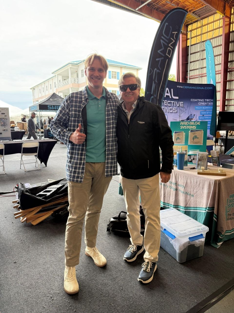 Two men beside an Arid Bilge Systems display at a boat show; the man on the left gives a thumbs-up.